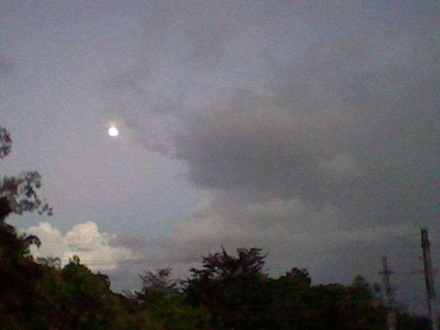 During sundown, the moon is visible in this image. Next to the distant moon, there is a grey cloud. Below, there are power lines and trees.