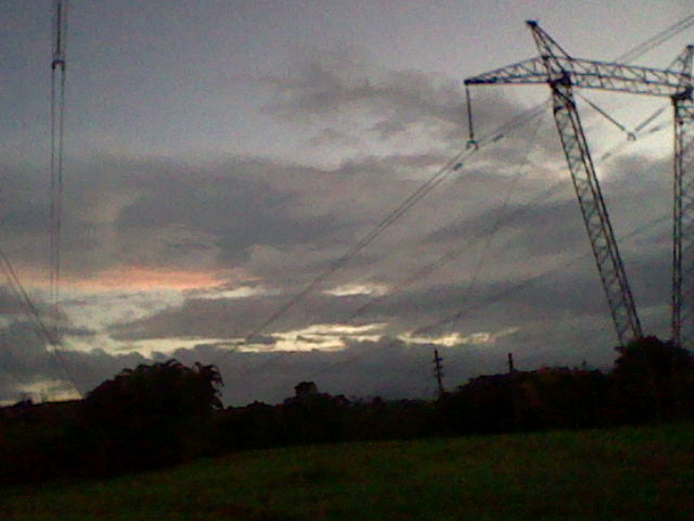 It's sundown and sky is cloudy. The ground is dim due to the camera settings. There are electricity lines above going towards the distance.