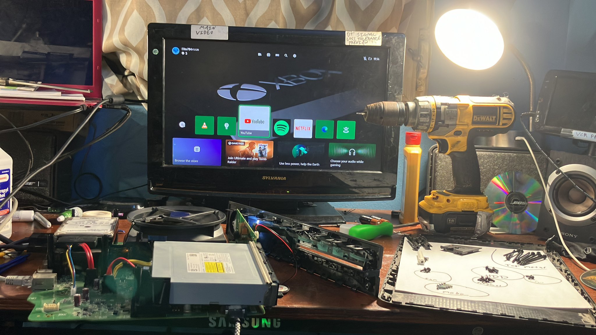 An XBox One without its plastic case on a table. There is a television at the back of the table displaying video from the XBox. To the left of the XBox there is a board holding screws and a Dewalt power tool.